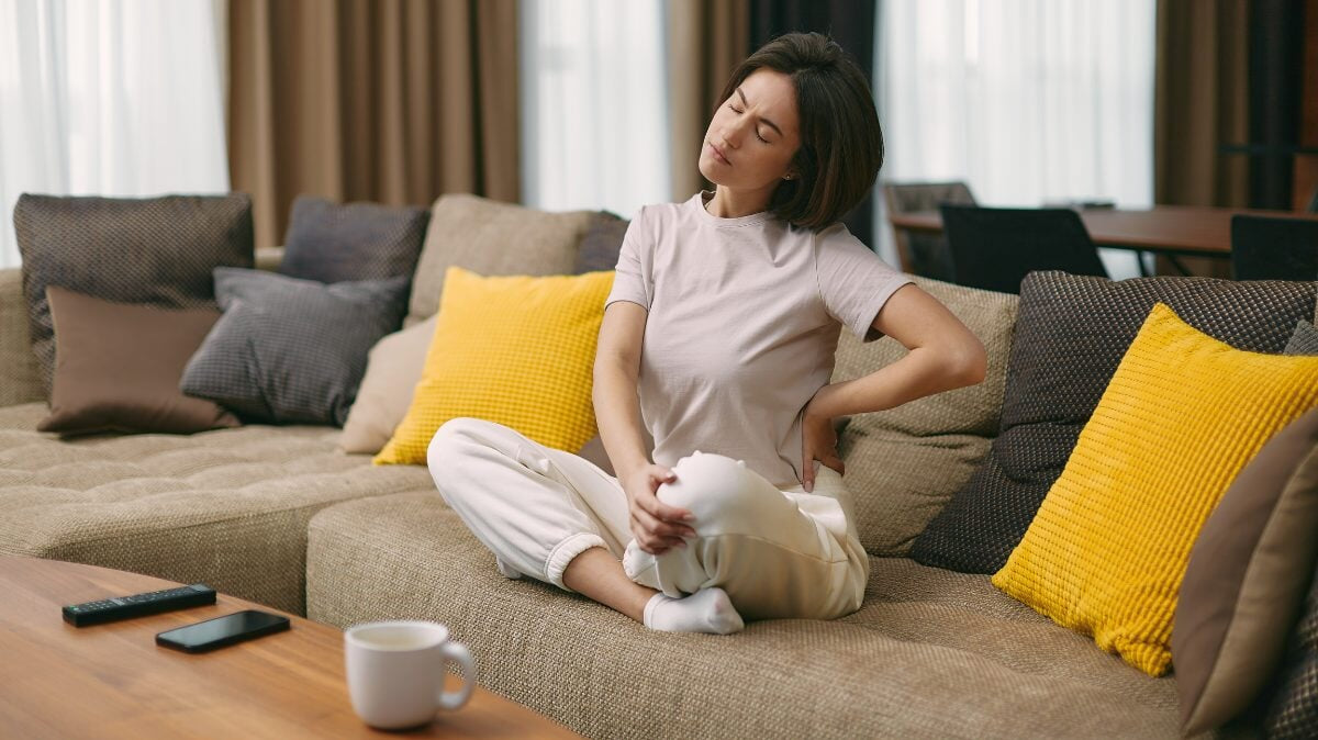 Une jeune fille fatiguée touchant le bas du dos ressentant un inconfort, souffrant d'une douleur soudaine due à un mode de vie sédentaire assise sur un canapé